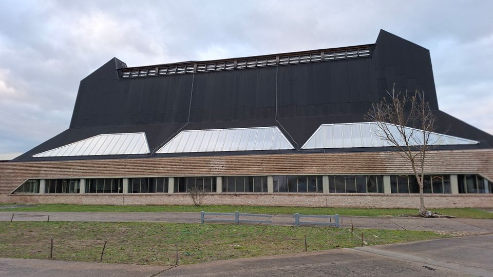 Ein modernes Gebäude mit einer schwarzen, steilen Dachstruktur, großen trapezförmigen Fenstern und einer unteren Fassade aus Holzverkleidung und Ziegeln. Im Vordergrund eine unbefestigte Wiese und ein Baum.