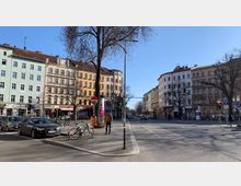 Straßenszene in einem urbanen Viertel mit mehrstöckigen Altbauten in verschiedenen Pastellfarben, Geschäften im Erdgeschoss und parkenden Autos sowie Fahrrädern am Straßenrand. Im Vordergrund stehen ein Baum, ein Mülleimer und ein Fußgänger, während die Straße im Hintergrund von Lichtsignalen geregelt wird.