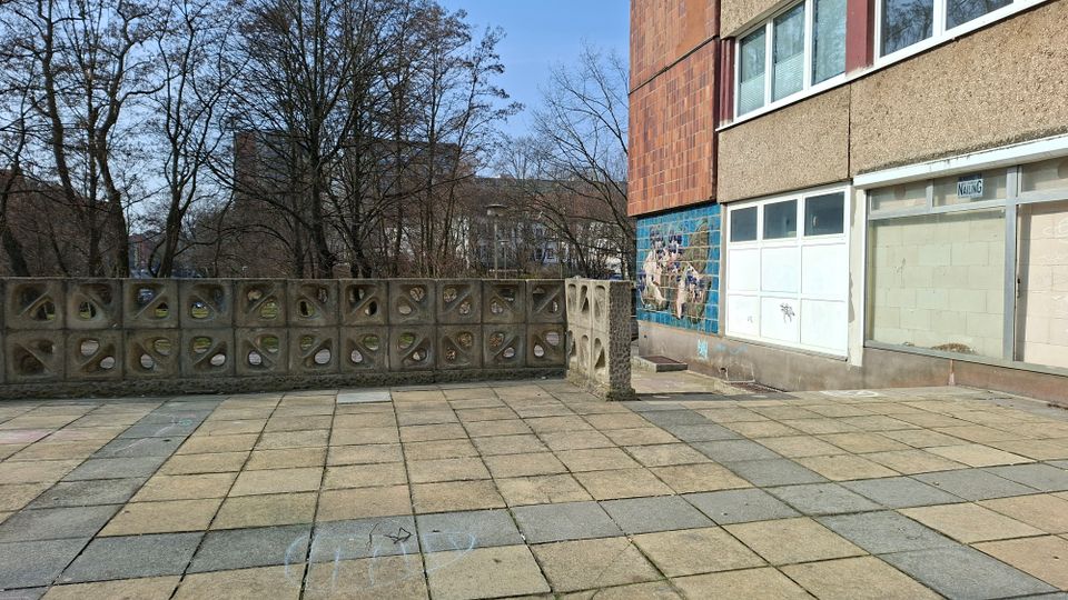 Gepflasterter Vorplatz neben einem mehrstöckigen Gebäude mit Beton- und Klinkerfassade, großen Fensterfronten und einem Keramikmosaik an der Wand. Eine niedrige Beton-Brüstungsmauer mit ornamentalen Öffnungen begrenzt die Fläche; dahinter stehen kahle Bäume und weitere Gebäude im Hintergrund.