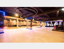 Großer Innenraum mit modernem Club-Design, heller gefliester Boden, verspiegelte Säulen, abgehängte schwarze Decke mit sichtbaren Rohren und kreisförmigen Leuchtelementen. Im Hintergrund Sitzbereiche mit goldenen Dekorelementen und großen Fenstern mit dunklen Vorhängen.