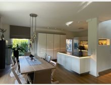 Moderne Wohnküche mit offenem Grundriss, holzfarbenem Esstisch und beige gepolsterten Stühlen. Die Küche verfügt über weiße Hochglanz-Schrankfronten, eine beleuchtete Kücheninsel und eine Glastür mit Blick ins Freie; Pflanzen und Dekorationen ergänzen den Raum.