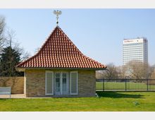 Ein kleines Gebäude aus gelben Backsteinen mit rotem Ziegeldach und einem goldenen Adler auf der Dachspitze steht auf einer Rasenfläche, umgeben von einem Zaun. Im Hintergrund ist ein Hochhaus mit der Aufschrift „hotel Mercure“ zu sehen.