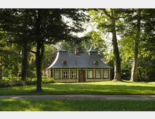 Kleines, historisches Gebäude mit gelber Fassade, schwarzen Verzierungen und geschwungenem Schieferdach, umgeben von hohen Bäumen und einer Rasenfläche in einem parkähnlichen Umfeld.