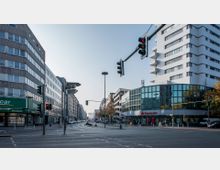 Straßenszene in einer städtischen Umgebung mit mehrstöckigen Büro- und Wohngebäuden. Im Vordergrund ist eine Straßenkreuzung mit Ampeln, während moderne Glasfassaden und Schilder, darunter eine Santander-Bank und eine Autovermietung, sichtbar sind.