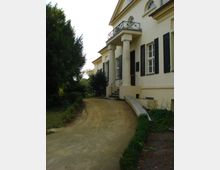 Seitenansicht eines historischen Gebäudes mit cremefarbener Fassade, schwarzem Fensterladen und einem Säuleneingang mit Balkon darüber. Ein geschwungener, sandfarbener Weg führt durch eine gepflegte Gartenanlage mit Hecken und Bäumen.