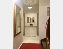 Ein Flur in einem Wohngebäude mit weiß gestrichenen Wänden und einem roten Teppichläufer auf dem Boden. Im Hintergrund steht ein weißer Schrank mit einer Blumenvase, darüber ein rechteckiger, goldgerahmter Spiegel; links hängt ein weiterer goldgerahmter Spiegel, rechts ist ein Treppengeländer aus Holz sichtbar.