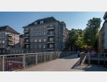 Wohngebäude aus der Gründerzeit mit grauer Fassade, mehreren Balkonen und weißen Fensterrahmen, im Vordergrund eine Fußgängerbrücke mit Metallgeländer. Rechts daneben stehen Bäume, die teilweise Schatten auf die Umgebung werfen.