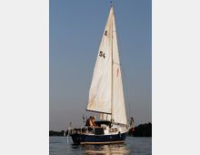 Segelboot mit weißem Segel und dunkelblauem Rumpf auf einem ruhigen Gewässer, bei klarem Himmel. Eine Person befindet sich an Deck, im Hintergrund sind leicht bewaldete Ufer und andere Segelboote erkennbar.