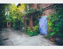 Hinterhof mit einem roten Mauergebäude, teils bewachsen mit Efeu und Pflanzen. Eine blaue Tür ist sichtbar, umgeben von Sträuchern und Bäumen auf einem Betongrund.