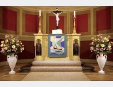 Altarraum einer Kirche mit rotem Hintergrund und goldfarbenen, verzierten Elementen. In der Mitte steht ein Kreuz mit einer Jesusfigur über einer Bibel; darunter hängt ein blau-grünes Wandtuch mit einem Segelschiff-Motiv. Links und rechts vom Altar befinden sich jeweils eine Statue und ein Blumenarrangement in großen weißen Vasen.