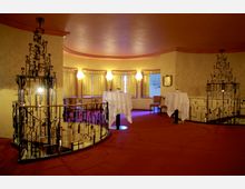 Innenraum eines eleganten Veranstaltungsraums mit roten Teppichböden, beigefarbenen Wänden und großen Fenstern mit goldenen Vorhängen. Im Vordergrund sind schmiedeeiserne Geländer um Treppenabsätze sowie mehrere hohe Tische mit weißen Tischdecken zu sehen.