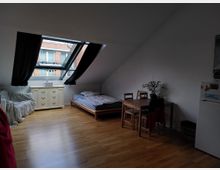 Ein Dachzimmer mit Dachfenster, durch das Außenfassaden eines Backsteingebäudes sichtbar sind. Der Raum enthält ein Bett, einen Esstisch mit Stühlen, eine Kommode und eine Couch, mit einem hellen Holzfußboden und schräger Decke.