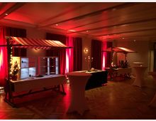 Ein Veranstaltungsraum mit Holzfußboden, rot beleuchteten Wänden und langen, roten Vorhängen. Im Raum stehen mit rot-weiß gestreiften Markisen dekorierte Verkaufsstände, weiße Stehtische und festliche Beleuchtung.