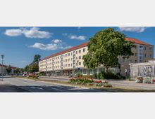 Breite Straße mit einem mehrstöckigen Wohn- und Geschäftshaus im Hintergrund, das eine beige Fassade und ein rotes Satteldach hat. Grünflächen mit Blumenbeeten und ein großer Baum säumen den Straßenrand, bei klarem Himmel und Sonnenschein.