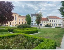 Städtischer Platz mit gepflegten Grünanlagen, niedrigen Hecken und Bäumen im Vordergrund. Im Hintergrund stehen historische, pastellfarbene Gebäude mit roten Ziegeldächern, darunter ein hellbeiges Gebäude mit großen Fenstern im klassischen Stil.