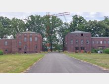 Zwei symmetrische Backsteingebäude mit rechteckigen Fenstern auf einer gepflasterten Straße, umgeben von Rasenflächen und Bäumen; im Hintergrund sind Teile einer großen Stahlkonstruktion, möglicherweise eines Sendemasts, sichtbar.