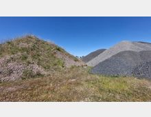 Zwei große Schotterhügel in einer offenen Landschaft; der linke Hügel ist teilweise mit Gras und kleinen Pflanzen bewachsen, während der rechte aus grauem, freiliegendem Kies besteht. Im Vordergrund ist eine Wiese mit niedriger Vegetation, dahinter ein klarer blauer Himmel.