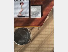 Top-down view of a wooden coffee table on a woven rug, holding a magazine and a small printed quote card. A round black ashtray or tray sits on the rug beside the table, with sunlight casting shadows across the floor.