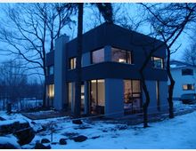 Modernes zweistöckiges Haus mit dunkler Fassade und großen Fenstern, durch die ein beleuchteter Weihnachtsbaum sichtbar ist. Das Gebäude steht in einer winterlichen Umgebung mit schneebedecktem Boden und kahlen Bäumen.