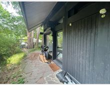 Außenansicht eines modernen Gebäudes mit dunkler Holzfassade und großen Fenstern, umgeben von Bäumen und grünem Garten. Ein schmaler Steinweg führt an der Hauswand vorbei, und vor dem Fenster steht ein Motorrad geparkt.