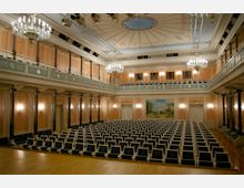 Eleganter Theatersaal mit einer Bühne im Vordergrund, Reihen gepolsterter Sitze und umlaufenden Galerien. Der Raum ist in Pastelltönen gestaltet, mit Säulen, kunstvoll verzierten Wänden, Kronleuchtern und einer Decke mit Malereien sowie einem großen Wandgemälde im Hintergrund.