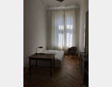 Ein schlichtes Schlafzimmer in einem Altbau mit hohen Decken und Stuckverzierungen. Der Raum enthält ein Bett, einen Holzschreibtisch mit Lampe, einen geflochtenen Stuhl und einen Schrank; durch ein großes Fenster mit weißen Vorhängen fällt Licht auf den Fischgrät-Holzboden.