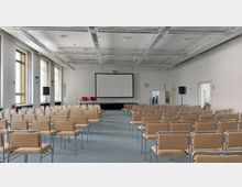 Großer Konferenzraum mit mehreren Reihen von beigefarbenen Stühlen, einer Bühne mit zwei roten Stühlen und einem weißen Projektscreen an der Rückwand. An den Seiten des Raumes befinden sich große Fenster mit vertikalen Lamellen und zwei Lautsprecher.
