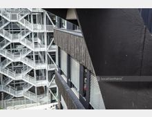 Blick auf die Außenfassade eines modernen Gebäudes mit Glas- und Metalltreppenhäusern, die sich in verschachtelter Form erstrecken, sowie einer angrenzenden Wand mit dunklem Holzverkleidung und Fenstern mit Jalousien. Im Vordergrund ist eine schwarze Stahlkonstruktion sichtbar.