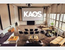 Ein heller Raum mit industriellem Charme, ausgestattet als kleines Kino mit einer großen Leinwand, auf der "KAOS Kino" steht. Es gibt eine Mischung aus Stühlen, Sofas, Teppichen und kleinen Tischen, sowie große Fenster, die viel Tageslicht hereinlassen.