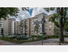 Mehrstöckiges Wohngebäude mit heller Klinkerfassade, umgeben von Bäumen und parkenden Autos auf einem Parkplatz. Im Vordergrund Straßenschilder mit den Namen „Brentanostraße“ und „Buggestraße“ sowie eine Bushaltestelle.