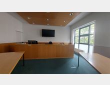 Ein kleiner Gerichtssaal mit einem erhöhten Richterpult aus Holz, einem TV-Bildschirm an der Wand, einem großen Fenster mit Blick auf Bäume und mehreren Tischen aus hellem Holz. Der Boden ist mit grünem Teppich ausgelegt.