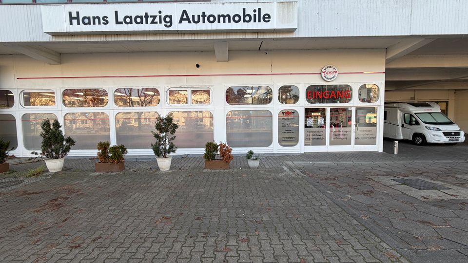 Außenansicht eines Autohauses mit der Beschriftung „Hans Laatzig Automobile“ auf einem weißen Schild. Große Glasfassade mit abgerundeten Fenstern, mehreren Topfpflanzen auf einem gepflasterten Vorplatz und ein Wohnmobil auf der rechten Seite unter einem überdachten Bereich.
