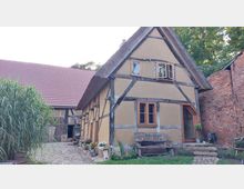 Fachwerkhaus mit Strohdach in einem ländlichen Hof, umgeben von Backsteinmauern. Im Vordergrund ist eine mit Pflastersteinen belegte Fläche sowie dekorativer Blumenschmuck zu sehen, daneben steht eine Holzbank mit einem Schild „Speicher anno 1554“.