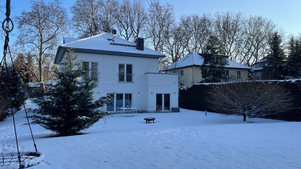 Blick auf den verschneiten Garten hinter einem modernen, weißen Einfamilienhaus mit flachem Satteldach und großen Terrassentüren. Im Vordergrund liegen eine schneebedeckte Rasenfläche mit Feuerschale und Tisch, umgeben von Hecken, Nadelbäumen und kahlen Laubbäumen unter blauem Himmel.