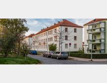 Wohnsiedlung mit zweistöckigen Gebäuden in einer ruhigen Straße. Links ein beige-weißes Mehrfamilienhaus mit roten Ziegeldächern und rechts ein Gebäude mit grün-weißen Balkonen; geparkte Autos und Bäume säumen die gepflasterte Straße.