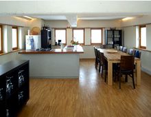 Moderne Wohnküche mit Holzfußboden, zentraler Kücheninsel mit Gasherd und Spüle, großem Esstisch mit mehreren Stühlen und Wandschränken, die Geschirr und Küchenutensilien enthalten. Die Fenster mit Holzrahmen lassen viel Tageslicht in den Raum.