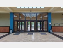 Eingangsbereich eines Gebäudes mit großen Glastüren und Fenstern, die in einen Außenbereich mit Bäumen führen. Der Innenraum hat helle Fliesen, Wände in Beige- und Blautönen, Holzelemente und eine Garderobe auf beiden Seiten.