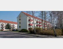 Mehrfamilienhäuser mit roten Satteldächern und roten Balkonen, umgeben von kahlen Bäumen, Büschen und einer gepflasterten Straße. Die Gebäude sind beige und drei Stockwerke hoch, mit kleinen Grünflächen davor.