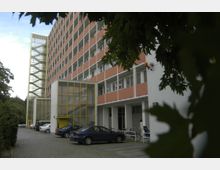 Mehrstöckiges Wohngebäude mit orangefarbenen und weißen Fassadenelementen, einer markanten gelben Treppe in einem verglasten Abschnitt und einem begrünten Gehweg mit geparkten Autos und Fahrrädern im Vordergrund. Links sind Bäume zu sehen.