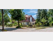 Ein Einfamilienhaus mit rotem Mansarddach und weißer Fassade, umgeben von einem gepflegten Garten und einem Metallzaun. Das Haus steht in einer ruhigen Wohngegend mit gepflasterten Straßen, Bäumen und grünem Bewuchs.