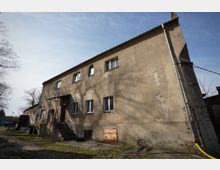 Zweistöckiges, längliches Wohn- oder Wirtschaftsgebäude mit verputzter, stellenweise abblätternder Fassade und mehreren rechteckigen Fenstern sowie einem kleinen überdachten Eingang mit Außentreppe. Vor dem Haus liegen ein gepflasterter Hof und Grasflächen, daneben stehen kahle Bäume unter blauem Himmel.