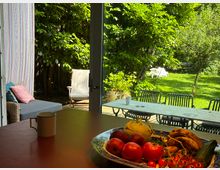 Blick aus einem Essbereich durch eine große Glastür auf eine Terrasse mit langem Tisch und Stühlen sowie einen grünen Garten mit Bäumen und Rasen. Im Vordergrund steht auf dem Tisch eine Schale mit Obst und Gemüse neben einer Tasse.