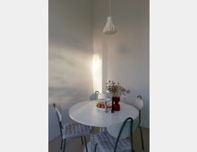 Heller, minimalistischer Essbereich mit rundem weißen Tisch, vier Stühlen mit gepolsterten Sitzkissen und einer weißen Pendelleuchte an hohen, weißen Wänden. Auf dem Tisch stehen eine Obstschale und eine rote Vase mit Blumen, seitliches Tageslicht wirft Schatten an die Wand.