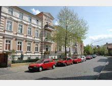 Straßenszene mit mehrstöckigen Wohnhäusern im Altbaustil auf der linken Seite, gekennzeichnet durch Ziegelmauern, weiße Stuckelemente und Balkone; davor stehen mehrere geparkte Autos, überwiegend in Rot, entlang einer gepflasterten Straße mit einem großen, belaubten Baum. Im Hintergrund sind weitere Gebäude und ein Baukran sichtbar.