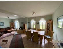 Wohnzimmer mit Essbereich in einem hellen Raum. Der Bereich ist möbliert mit einem Esstisch und sechs Stühlen aus Holz und Rattan, einer braunen Eckcouch, zwei Holzschränken, sowie einem kleinen Fernseher; helle, bodentiefe Fenster mit weißen Vorhängen lassen viel Tageslicht herein.