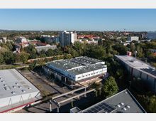 Luftaufnahme einer gemischten Stadtlandschaft mit einem Gewerbegebiet im Vordergrund, bestehend aus mehreren flachen Industriebauten mit weißen Fassaden und Flachdächern. Im Hintergrund sind Wohngebäude, viel Grün und ein vorbeifahrender Zug auf einer Bahnstrecke zu sehen.