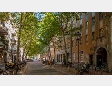 Eine ruhige Straße in der Stadt mit Kopfsteinpflaster, flankiert von mehrstöckigen Wohngebäuden mit hellen und bräunlichen Fassaden. Große Bäume spenden Schatten, während Fahrräder und eine Laterne den Straßenrand säumen.