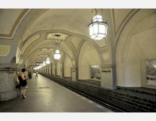 U-Bahn-Station mit hohen, hell erleuchteten Gewölbedecken und dekorativen Hängeleuchten. Auf dem Bahnsteig befinden sich Personen und an den Wänden sind historische Bilder angebracht, während der Schriftzug „Heidelberger Platz“ sichtbar ist.