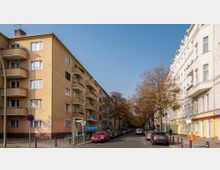 Straßenszene mit mehrstöckigen Wohngebäuden auf beiden Seiten; links ein gelbes Gebäude mit Balkonen, rechts ein weißes Haus. In der Mitte der Straße Autos, Bäume mit Herbstlaub und ein U-Bahn-Schild für die Station "Augsburger Straße".