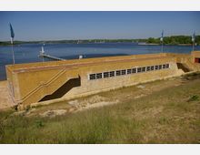 Gelbes Gebäude mit rechteckigen Fenstern und Außentreppen, gelegen an einem Ufer mit Blick auf einen See mit Steg und klarem blauen Himmel im Hintergrund. Im Vordergrund befindet sich eine grasbewachsene Fläche, und auf dem Gebäude wehen Fahnen.